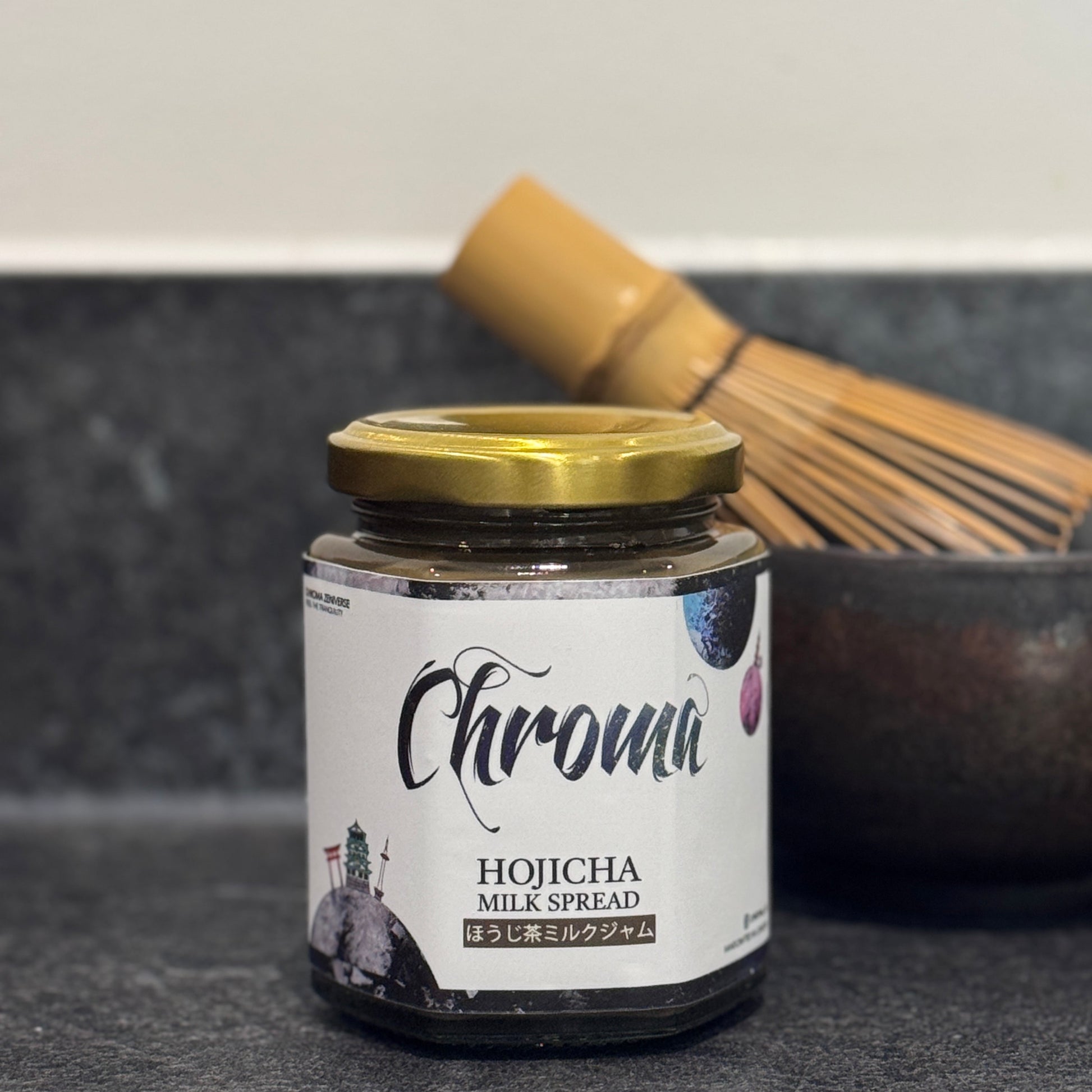 Jar of Chroma Hojicha Milk Spread on a dark surface with a whisk in the background