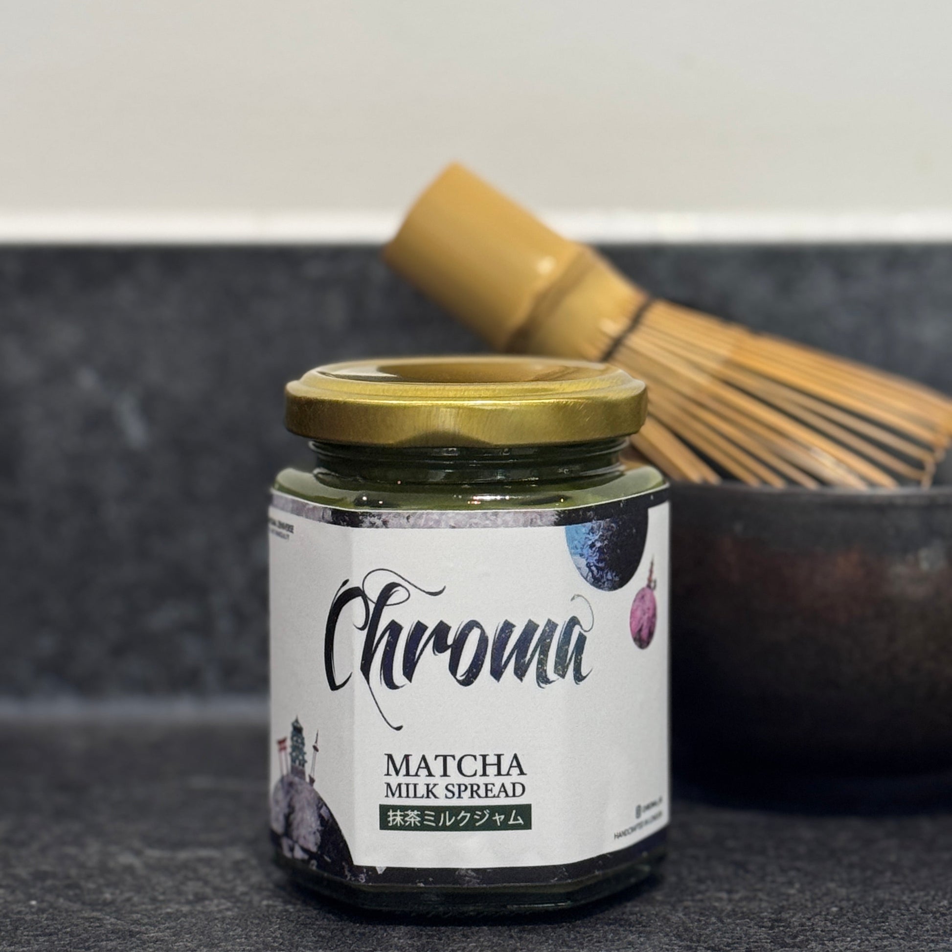 Jar of Chroma Matcha Milk Spread on a dark surface with a whisk in the background
